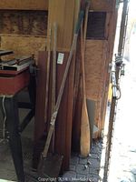 Photo showing wood boards, garden rake, shovel, and wooden paddles leaning against a wall in an outdoor area.