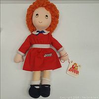 Front of the Little Orphan Annie plush doll showing red yarn hair, red dress with white collar, white belt, and black shoes. Doll holds original tag with Annie logo.