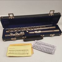 Disassembled silver Armstrong flute inside open black case with blue velvet lining, accompanied by sheet music.