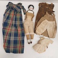 Full view of the china head doll with associated clothing items including a long plaid dress, brown dress, pantaloons, and an undergarment top.