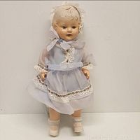 Full front view of the Fiberoid composition doll wearing a light blue sheer dress with lace and floral trim, with crocheted socks.