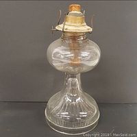 Full view front clear glass oil lamp with ribbed lower base and round upper reservoir, metal burner with wick on top.
