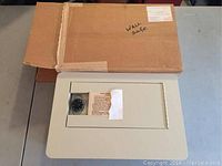 Top view of the beige Brookstone wall safe with visible combination dial lock and instruction label under protective window, with the safe partially out of its cardboard box.