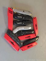 Top view of seven utility knives showing a mix of red and black handles, with two X-ACTO branded black knives and one silver ORCON knife.