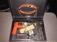 Open metal box with vintage orange and silver Black and Decker drill, orange cord, and various small accessories including drill bits and a clamp.