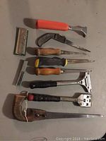Top-down photo of various hand tools including scrapers and saws with wooden and plastic handles on a grey surface.