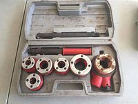 Top-down photo showing eight red and chrome pipe threading die heads and two handle extensions organized in grey plastic case