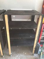 Full view of the wooden rolling shelving unit shows three slatted shelves with black finish, vertical supports in wood tone, caster wheels at the base. Light scuff marks visible on shelves.