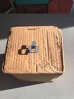 Closed cardboard box with wear marks, sealed, with single 1/2 inch plywood bracket placed on top for display.
