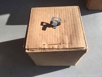 Top view of unopened cardboard box with a single metal plywood bracket placed on top for size reference