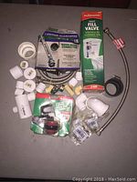 Overview photo of all plumbing items in lot, showing hoses, white PVC fittings and boxed Fluidmaster toilet parts.