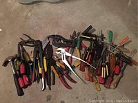 Wide view of assorted screwdrivers, pliers, hammer, and other hand tools on concrete floor with visible grease and wear.