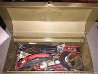 Photo of open olive green metal tool box showing assorted hand tools inside, including wire crimper, folding saw, pliers, tape, and screwdrivers.
