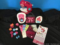 Red and white children's sewing machine with teal knob, fabric patterns, decorative patches, instruction pamphlet, and carrying case on black background.