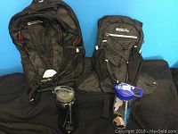 Two black Mountain Warehouse knapsacks with branded Force H2O hydration reservoir and two water bottles placed on black fabric background.