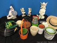 Photo showing garden hose coiled with nozzle, multiple flower pots in different colors, three fairy statues, and part of a garden clock.