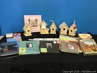 Photo of 5 unfinished wooden bird houses of various shapes and sizes along with 4 photo albums and puzzles arranged on a black tablecloth against a blue background.