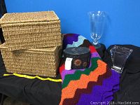 Photo showing complete lot with two woven baskets stacked, colorful crocheted afghan, black oval tole storage box, and two clear glass vases including a pedestal and rectangular shape.