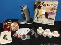 Philips Senseo coffee maker unit with power base, coffee pods, Reunion Island coffee pouches, 6 white espresso cups with saucers, and user manual.