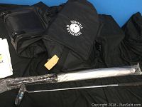 Black golf travel bag with wheels, side pockets, shoulder strap, and a towel seen on the left edge. A golf putter is partially wrapped in plastic lying in front.