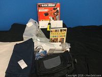 Full view of entire lot showing Black & Decker drill, box, handyman guide book, folded blue car blanket, tool pouch, and assorted items on black backdrop.