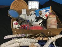 Photo showing mixed household items in a box including covered hangers, small mirror, electric iron, Coca Cola coasters, ice cube trays, glass cup, and ceramic vase