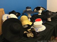 Full view of the 10 ladies hats arranged on a black surface against a blue background, showing variety of styles including felt, woven, and faux fur in black, white, leopard print, and yellow tones.