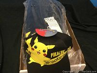Photo showing one black Pokemon cap featuring Pikachu character on front, packaging visible in background.