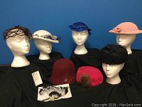 Group photo of eight vintage ladies hats arranged on mannequin heads and displayed on a black cloth against a blue background.