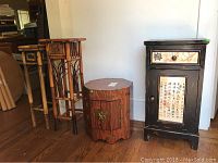 Wide photo showing two tall bamboo stands, one small scalloped wooden cabinet with brass latch, and one black wooden cabinet with drawer and door painted with Asian script.