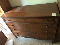 Full front view of carved wooden chest of drawers showing four drawers with rosette knob handles, veneer inlay on drawer fronts, and wooden top with visible scratches.
