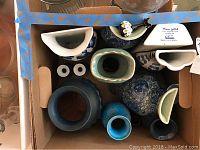 Top view of ten decorative items inside a cardboard box, showing varied shapes and colors including blue, white, green, and black ceramic and porcelain vases and bottles.