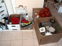 Wide photo showing multiple boxes containing kitchenware items including blender parts, red ceramic canisters, bakeware, and assorted glass containers.
