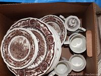 Top-down view of brown Americana Style House Ironstone plates, cups, and a lidded sugar bowl inside a cardboard box.