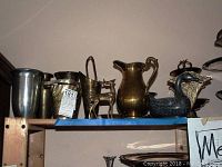 Shelf with assorted metal pitchers, vases, and decorative figurines including a tall silver-toned vase, brass deer, and bluish duck figurine.