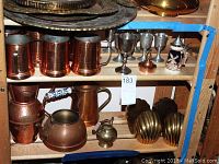 Wide view of copper mugs, teapots, brass decor, silver interior goblets and decorative stein on wooden shelf