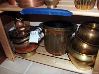 Photo showing multiple copper vessels arranged on a wooden shelf, highlighting variety of sizes and metallic tones.