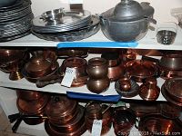 Copper and brass kitchenware items displayed on shelves, including tea kettle, saucepans, double boiler, and pedestal bowl. Items show wear and patina.