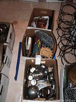 Three cardboard boxes containing assorted metal kitchenware, wooden handle flatware, trivets, and kitchen utensils.
