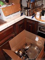 Wide view of stacked stainless steel cookware including pots and large fish poacher on counter and in cardboard box.