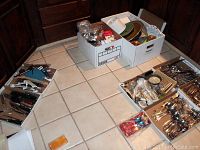 Photo shows several boxes containing mixed kitchen flatware, various utensils, and kitchen tools on tiled floor.