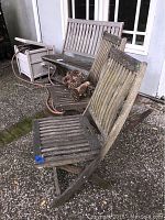 Three pieces of teak outdoor furniture: one bench and two chairs, all wooden with slatted seats and backs, placed outdoors on patio surface. Furniture has visible weathering and wear.