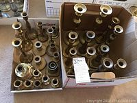 Overview of many brass candlesticks in two cardboard boxes showing wide variety of sizes and designs.