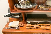 Flatware knives with black handles displayed on a wooden surface along with a silver plate covered container and a wooden twisted utensil set.