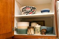 Wide view of shelf showing all pottery: Running Raven salad bowl, blue and white bowl, tall two-tone container, smaller textured pottery, sugar container, small cup, and bowl
