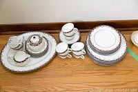 Photo showing full set of dinnerware arranged on floor, including cups, saucers, plates of various sizes, creamer, sugar bowl, and platter with distinct decorative borders.