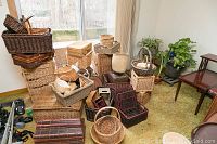 Overview of a collection of assorted woven baskets of various sizes and shapes stacked together near large window. Potted plants and small wooden table visible in the background.