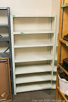 Full view of the white metal utility shelving unit with six shelves showing note writing on the bottom shelf.