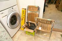 Two wood slatted folding chairs with metal frames leaning against a wall, cardboard box with BBQ tools beside them, washer and dryer visible nearby.