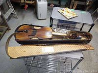 Violin with bow placed inside a broken case with damaged lining, showing significant wear and missing elements.